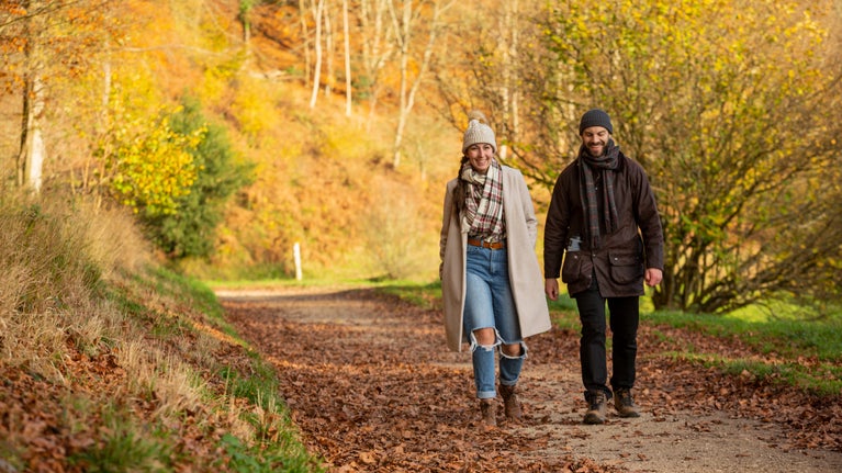 Man and woman in warm clothing walking along footpath covered with leaves, with autumn foliage all around